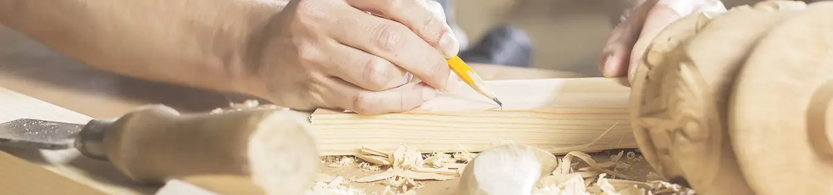 Hand zeichnet mit Bleistift eine Markierung auf eine Holzleiste, daneben Stechbeitel und Hobelspäne