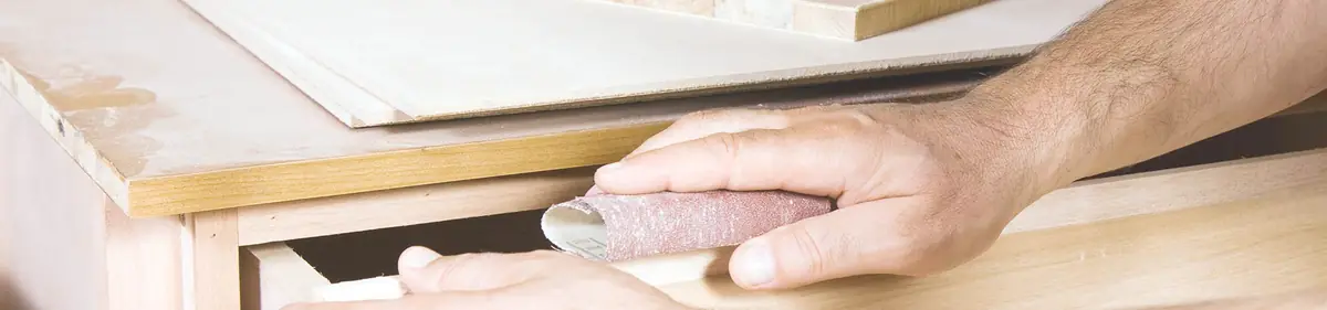 Hände schleifen mit Schleifpapier die Kante eines Möbelstücks